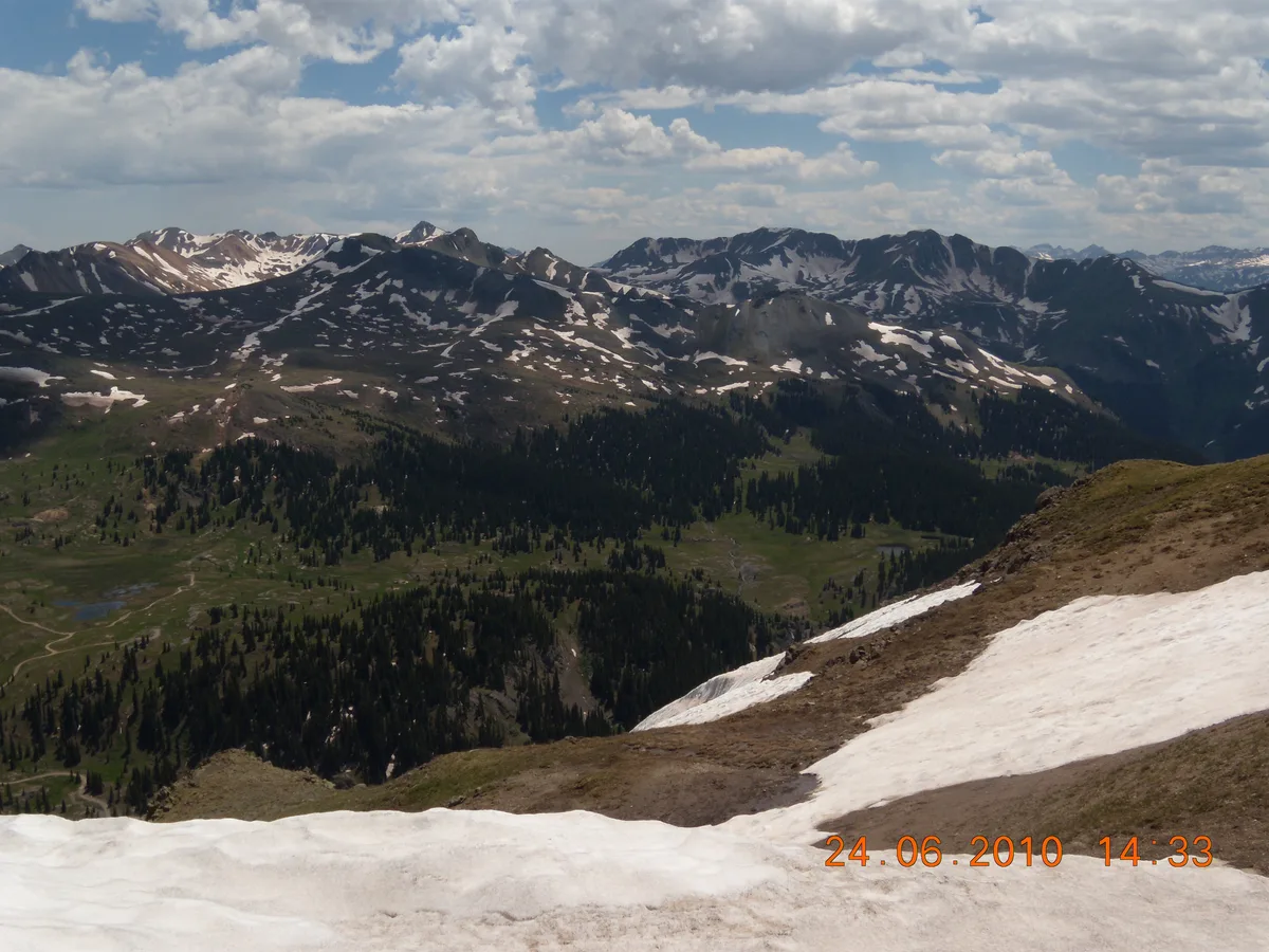 Wide view from a snowy alpine pass, with patchy snow in the foreground, dark evergreen forests below, and rugged, snow-streaked San Juan peaks stretching across the horizon under a partly cloudy sky. The scene feels high and open, with bright midday light and deep mountain valleys visible in the distance.