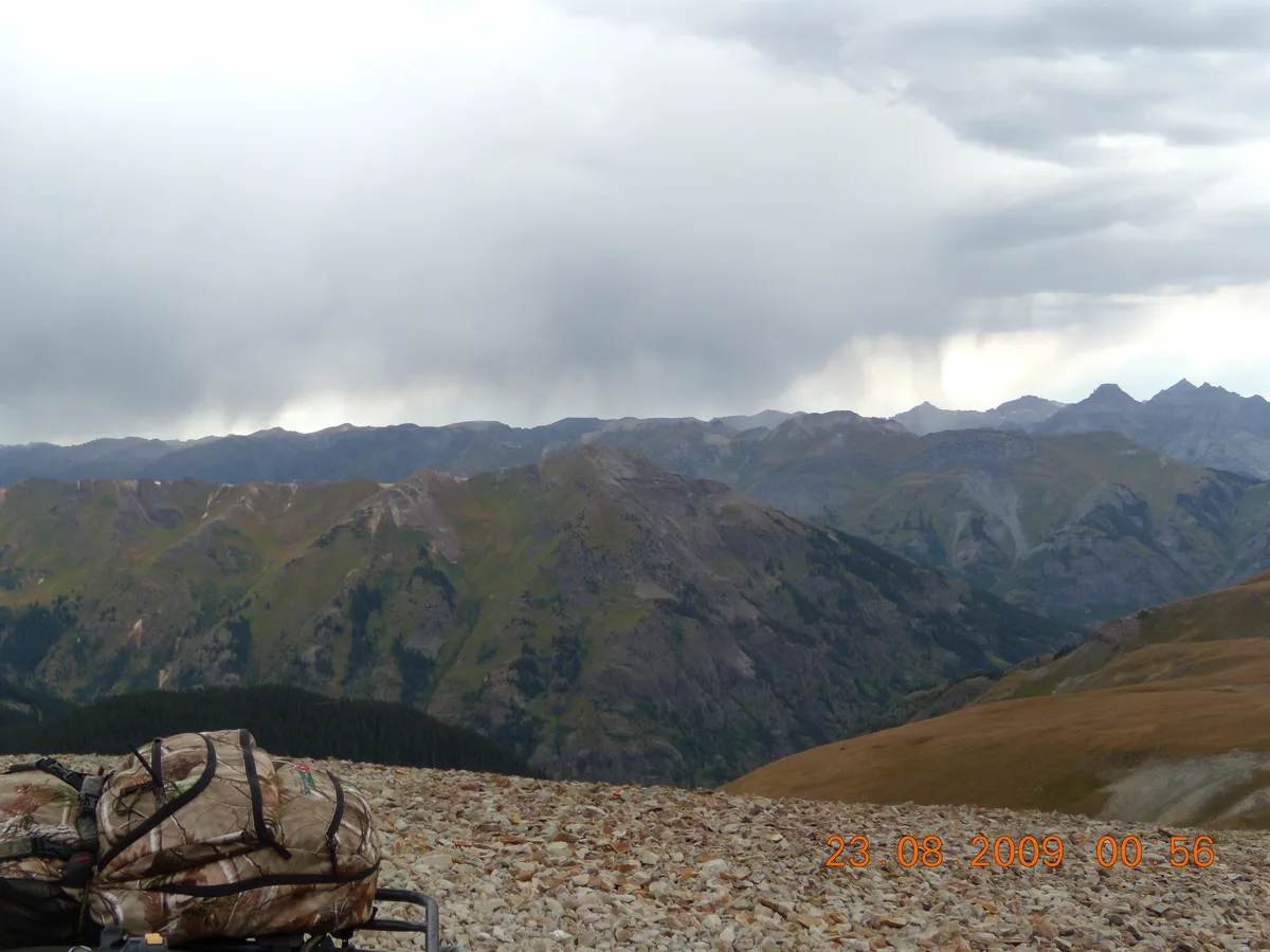 A rocky alpine overlook on Engineer Pass looks out over layered San Juan Mountain ridges under a heavy gray sky, with distant peaks fading into the haze. In the lower left, a camouflaged pack or gear bag sits on the rough gravel foreground, while a brown grassy slope drops away at right.