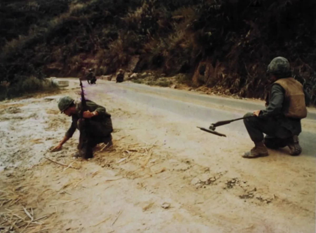 Two soldiers in camouflage kneel beside a narrow mountain road, sweeping the dirt shoulder with mine detectors while rifles are slung across their backs. The road curves into a steep, wooded hillside under dim, overcast light.