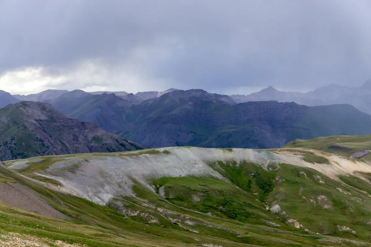 Wide view from a high alpine pass shows rolling green-and-gray tundra ridges dropping into dark, rugged San Juan Mountains under a heavy overcast sky. Distant peaks fade into mist, with soft morning light breaking through clouds on the left.