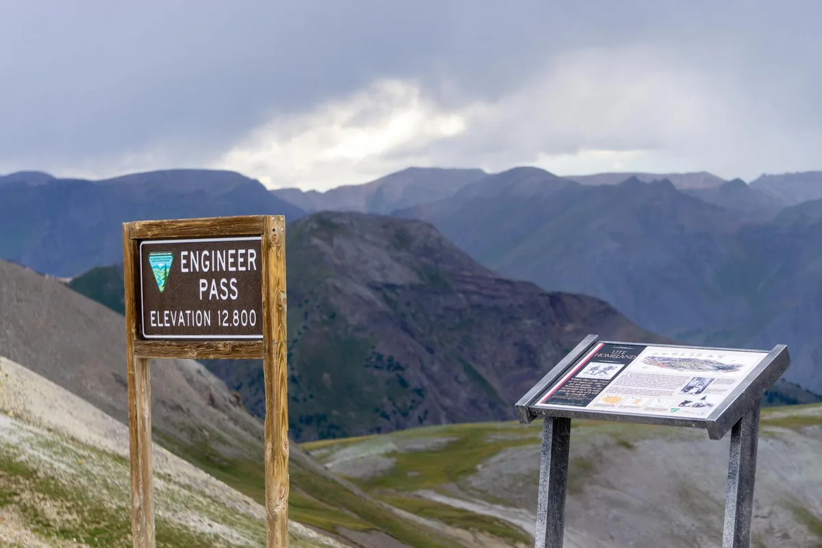 Engineer Pass trail sign and a nearby interpretive panel stand on a rocky alpine slope, with sweeping layers of dark blue mountain ridges fading into a cloudy morning sky. The foreground shows sparse tundra and gravel at 12,800 feet, emphasizing the high pass and vast 360-degree view.