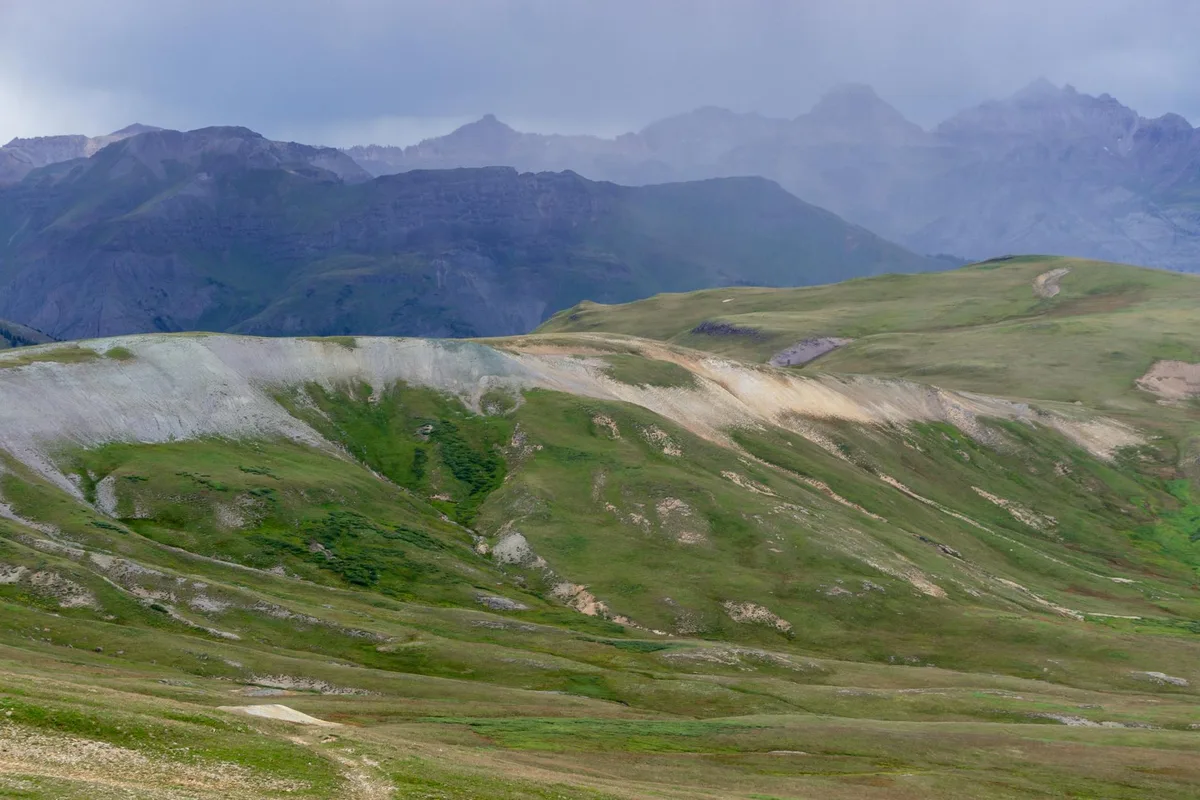 Rolling green alpine slopes and pale scree ridges stretch across the foreground, leading to a hazy line of rugged San Juan peaks under an overcast morning sky. The wide view feels high and open, with soft light flattening the distant mountains into layered blue-gray silhouettes.