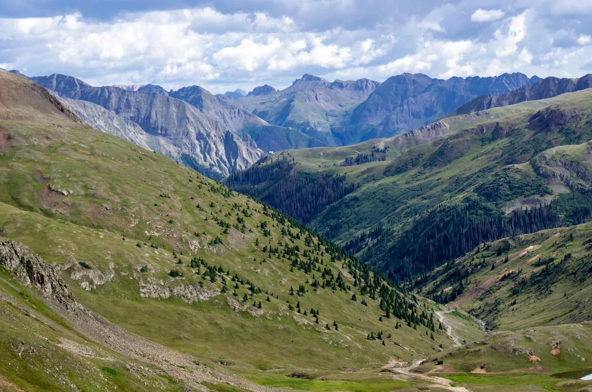 Sweeping view from a high alpine pass over broad green tundra slopes dotted with small evergreens, dropping into a deep valley framed by rugged gray peaks. Puffy clouds cast soft light across the layered San Juan Mountains in the distance.