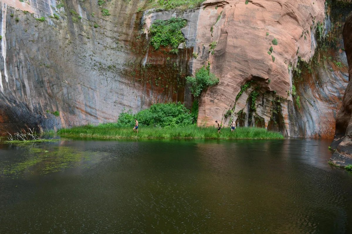 A calm pool reflects steep red and gray sandstone walls streaked with dark mineral stains, with a narrow strip of bright green grass and shrubs along the far bank. Three small people stand beneath the canyon face, emphasizing the scale of the tall rock amphitheater.
