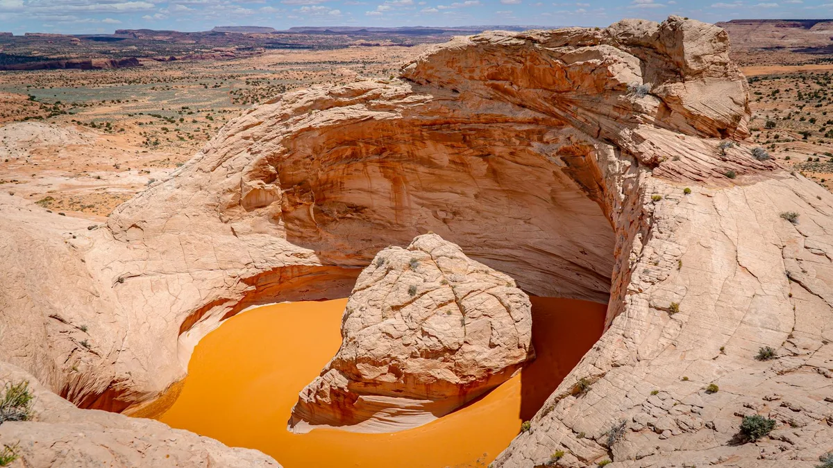A rounded sandstone pothole filled with bright orange water sits in the middle of a pale, wind-sculpted rock basin, with layered canyon country stretching into the distance under a clear blue sky. Sparse desert shrubs dot the sunlit rock, and the warm golden light brings out the red and tan bands in the stone.
