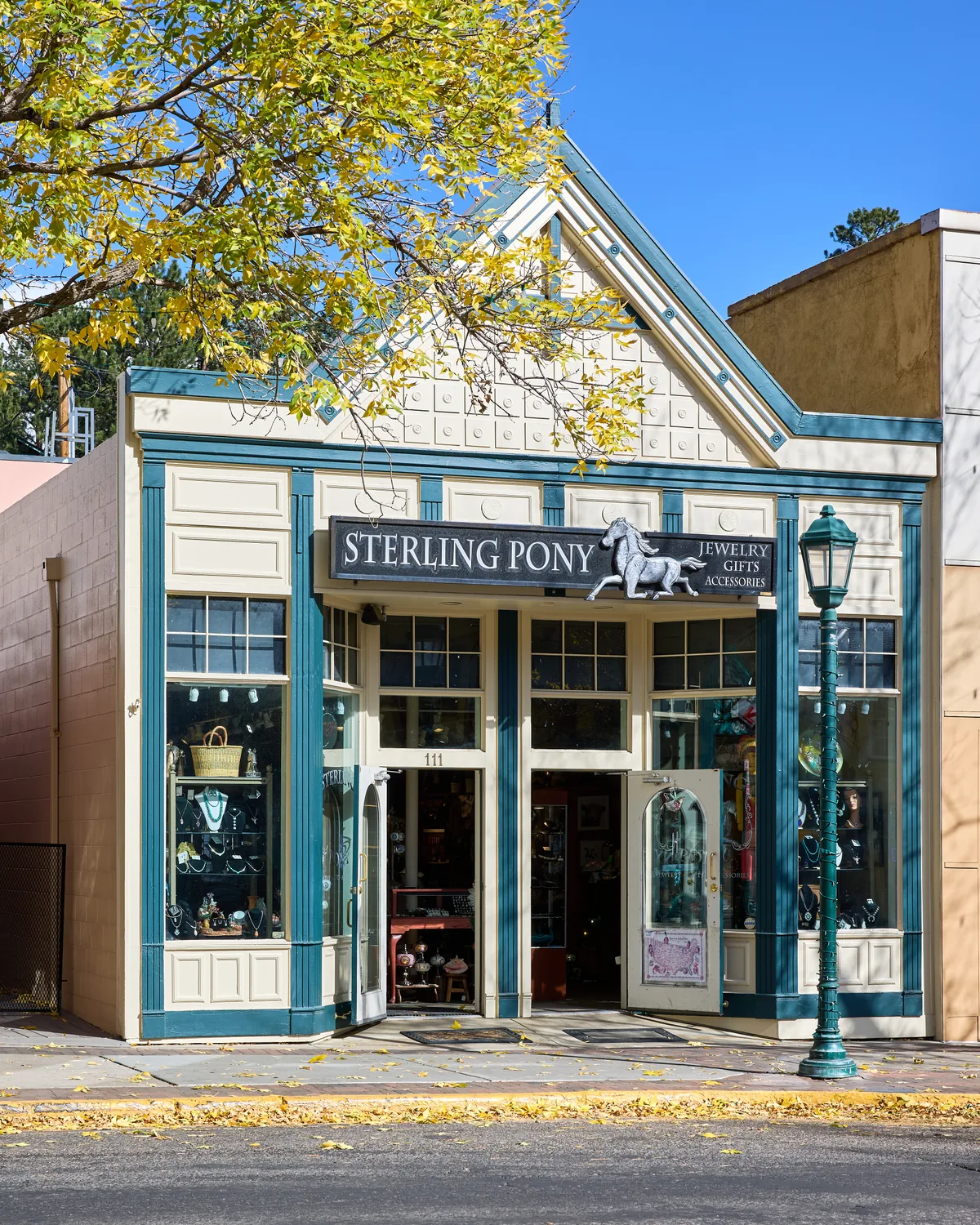 A quaint downtown Estes Park storefront for Sterling Pony fills the frame, with teal trim, cream paneling, and jewelry displays visible through the windows. Golden autumn leaves hang across the top edge under a clear blue sky, while a green streetlamp stands beside the open double doors.