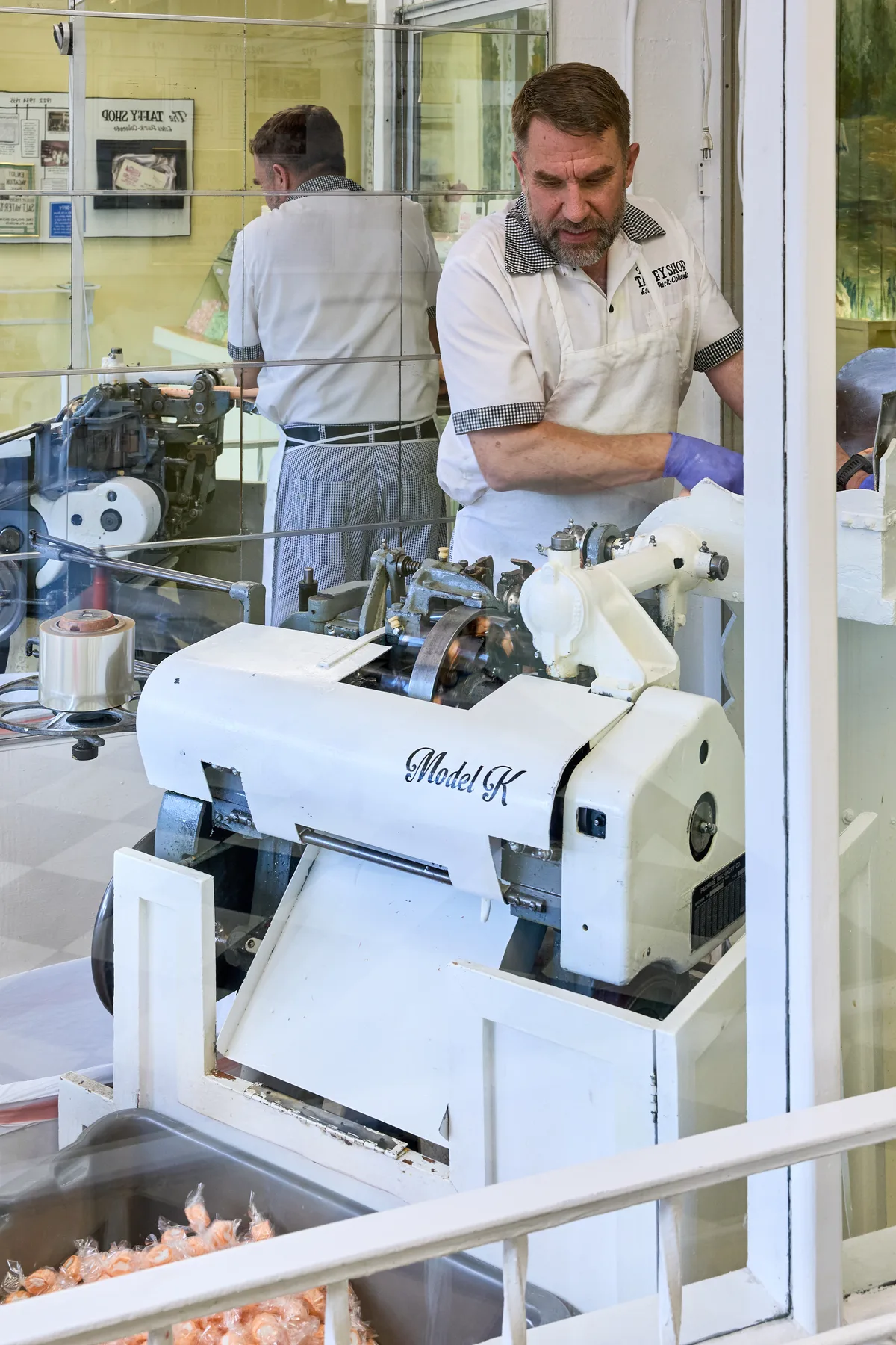 Inside a candy shop window, a man in a white apron and purple glove works a white Model K machine as wrapped taffy pieces pile up below. His reflection and the shop’s yellow interior walls show through the glass, giving the scene a bright, hands-on factory feel.