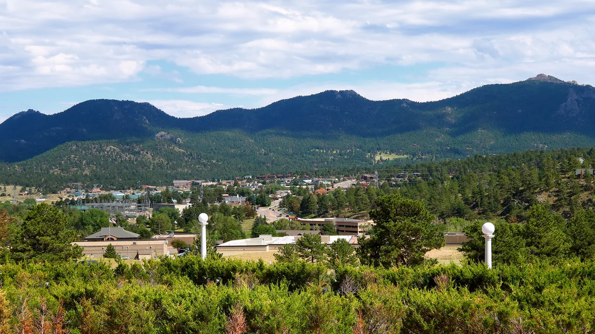 Wide view over Estes Park with downtown buildings nestled among evergreens below a broad, dark-green mountain ridge. In the foreground, trimmed shrubs and white lampposts frame the town under a bright, partly cloudy sky.