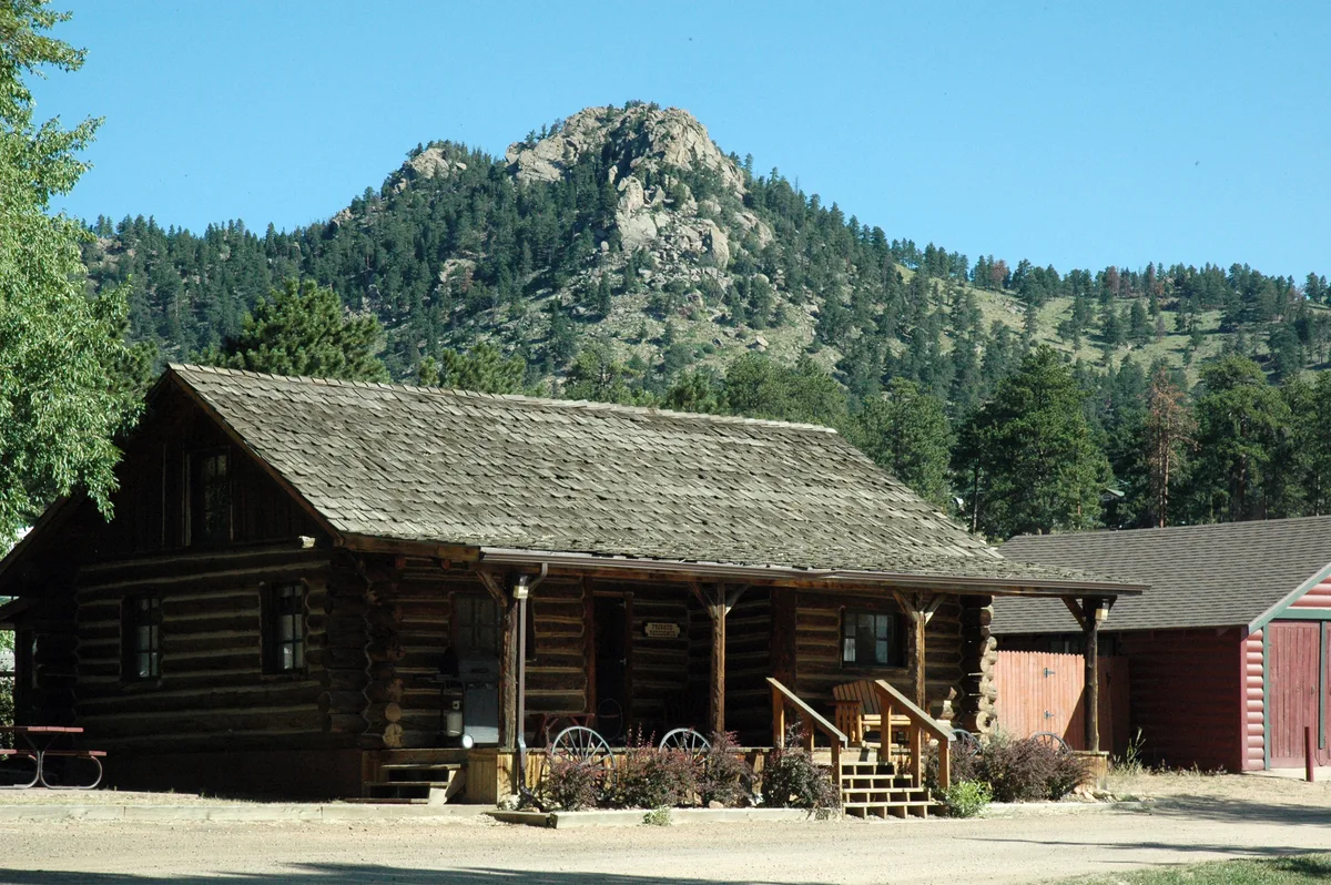 A rustic log cabin lodge sits in bright daylight beneath a steep, pine-covered mountain ridge, with a weathered shingle roof and small front porch in the foreground. Clear blue sky and sunlit trees frame the scene, giving it a quiet mountain-town feel.