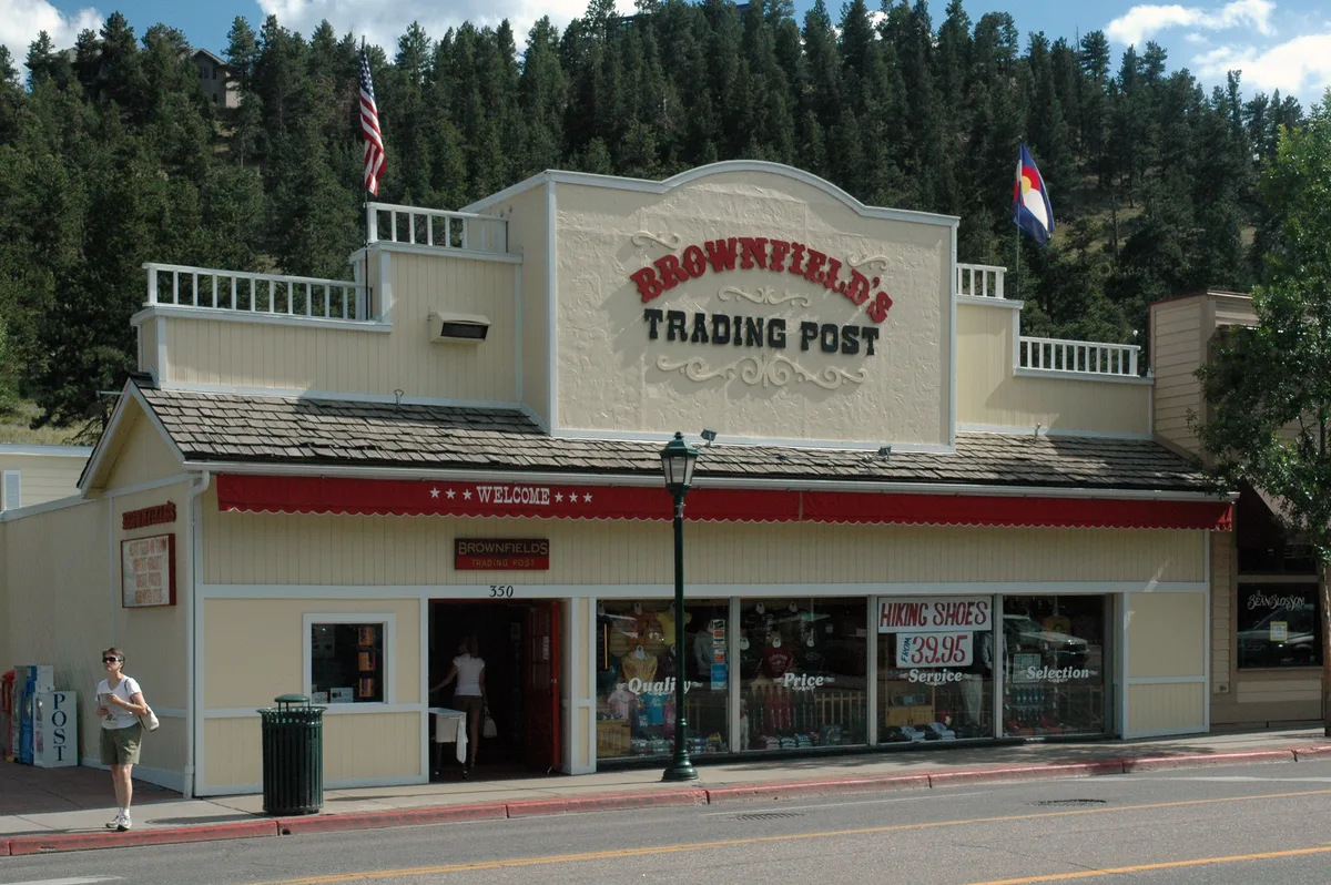 Brownfield’s Trading Post sits along Elkhorn Avenue in a cream-colored Western storefront with a red awning, large display windows, and flags flying above the roofline. A pedestrian walks past the curb in front, with a steep pine-covered hillside rising behind the downtown street.