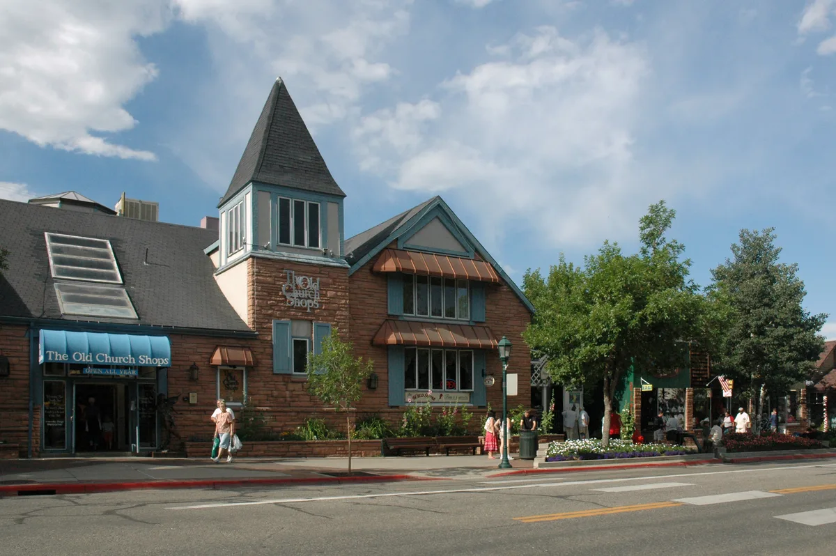 The Old Church Shops line Elkhorn Avenue in downtown Estes Park, with a pointed tower, blue awnings, and brown shingle siding under a bright, partly cloudy sky. People stroll along the sidewalk past street trees, flower beds, and shopfronts in warm late-day light.