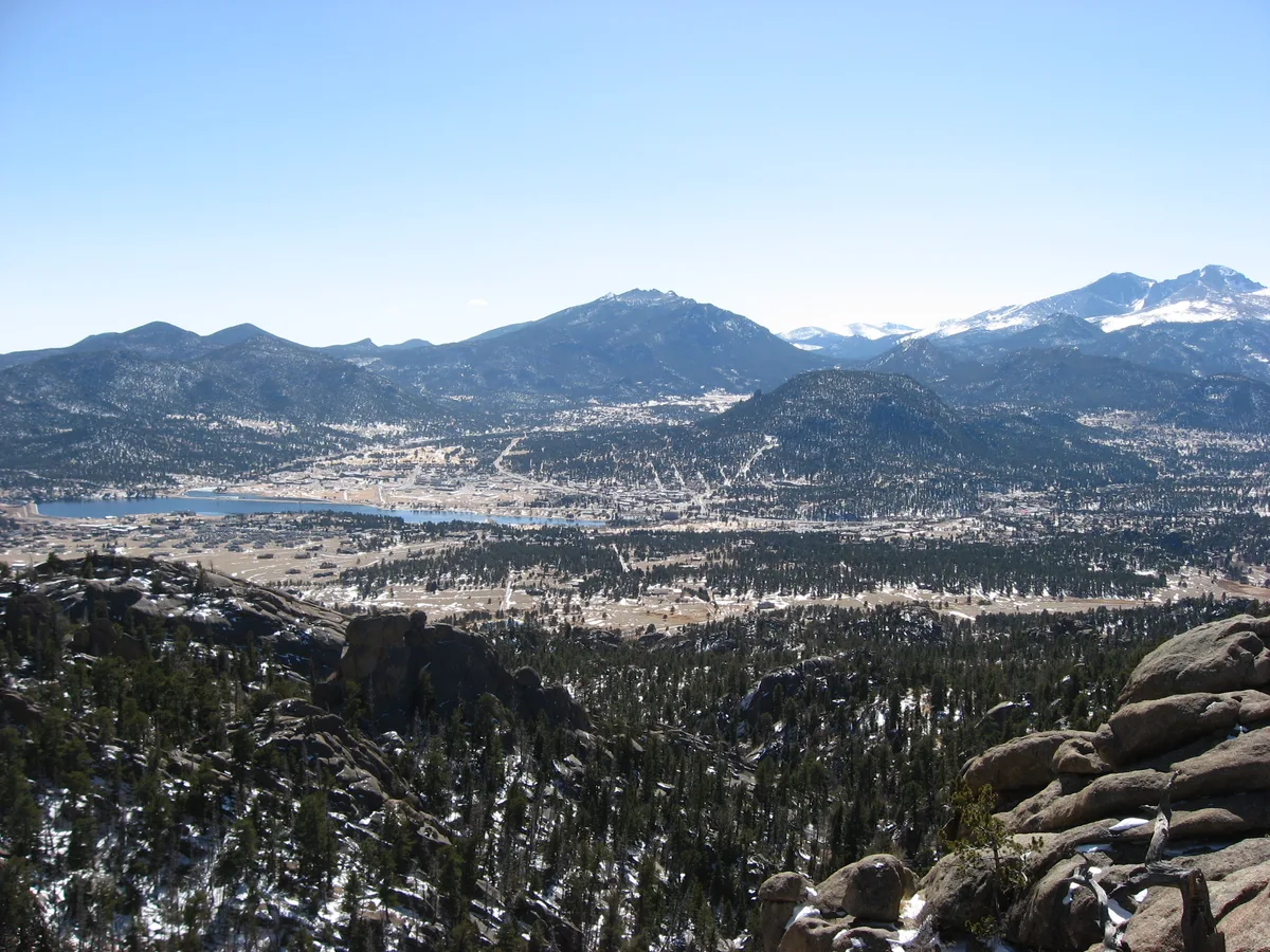 A sweeping view from a rocky mountain overlook shows Estes Park spread across the valley below, with the lake and town threading through dark evergreen forests. Snow-dusted peaks rise in the distance under a clear blue sky, while sunlit boulders frame the foreground.