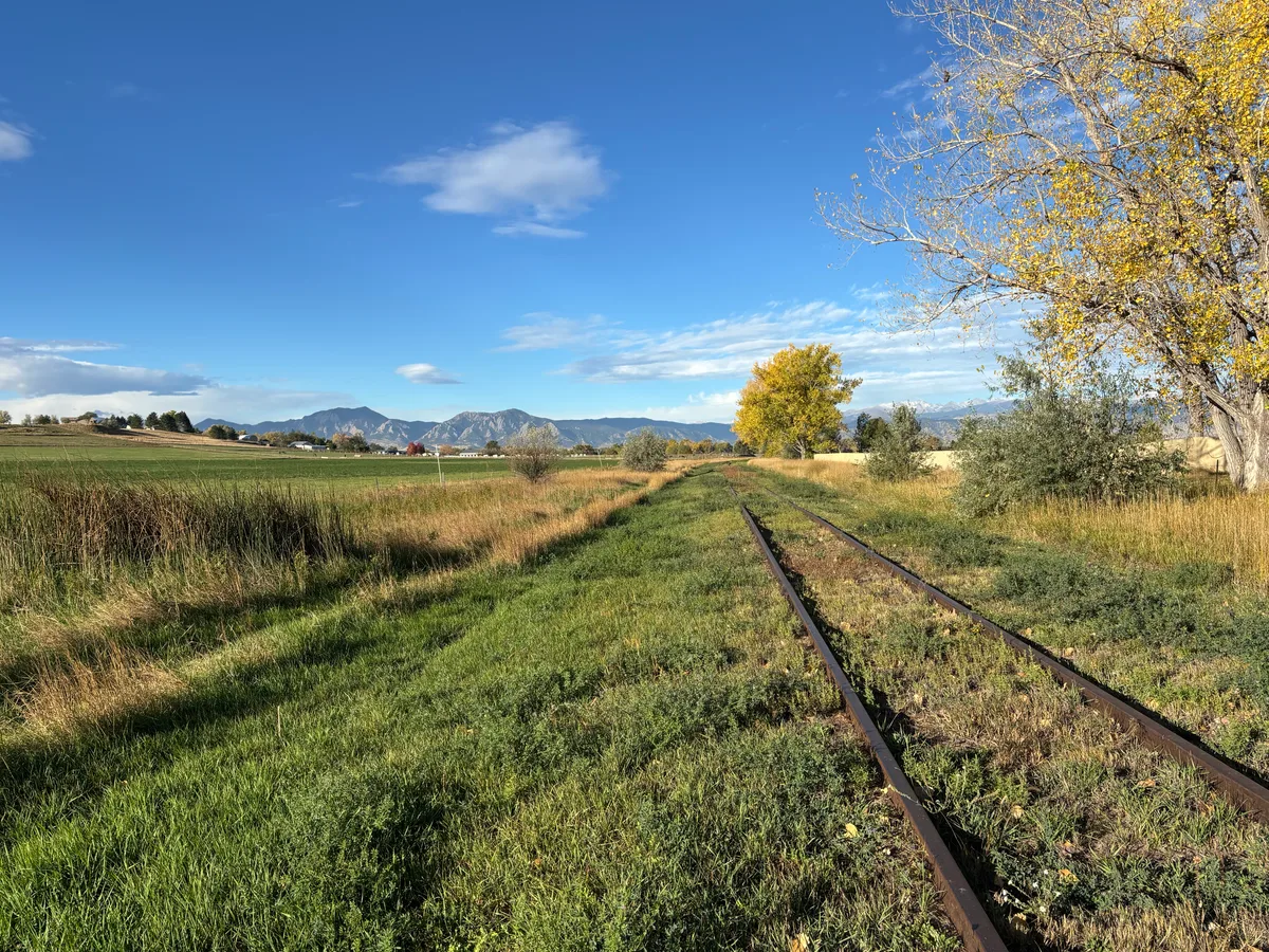 Rusty railroad tracks cut through a grassy field toward the Flatirons, which rise in a hazy line across the horizon under a clear blue sky. Golden autumn trees frame the scene, with long late-day light warming the grass and foothills.