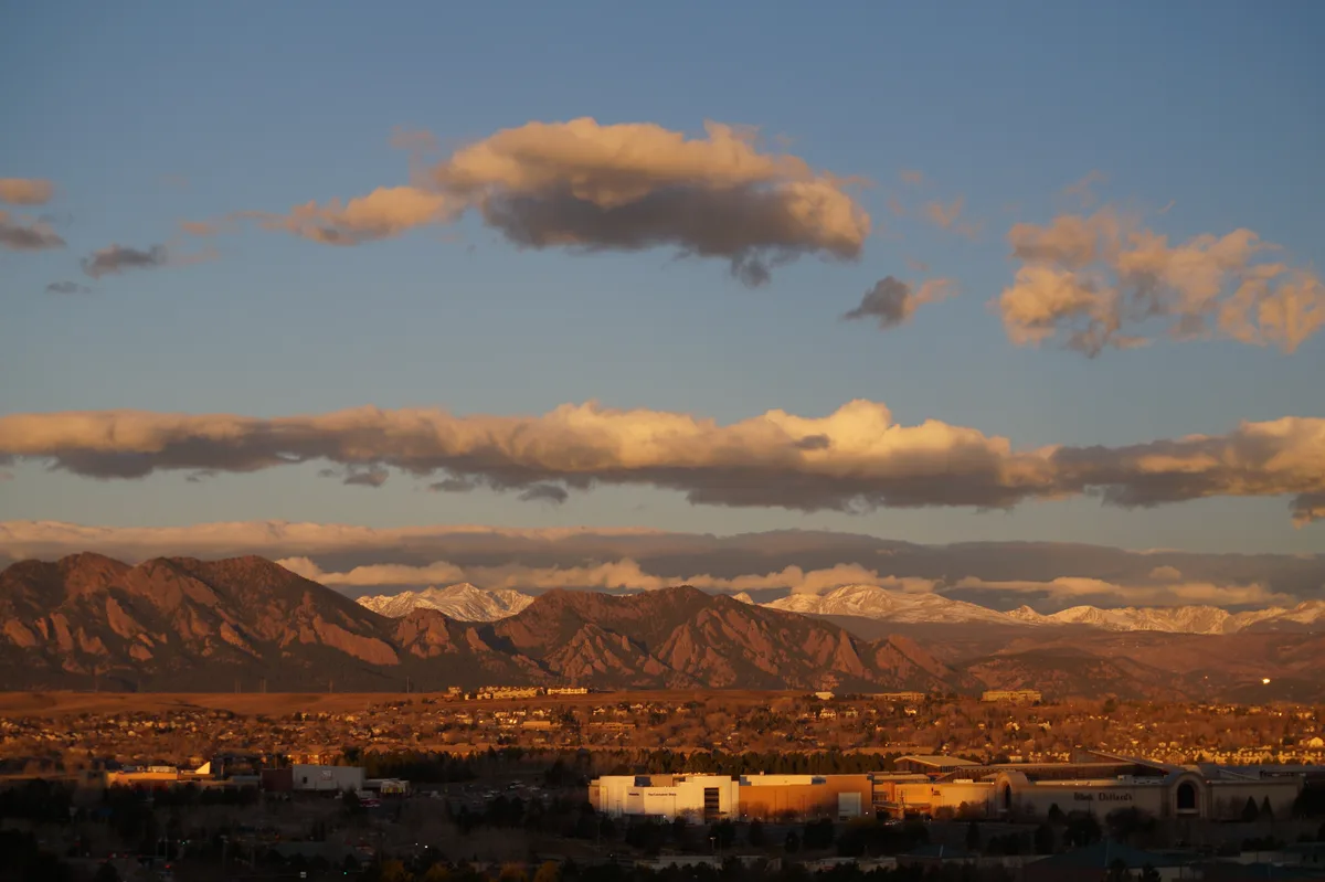 Golden-hour light warms the Flatirons and nearby foothills, with the snow-dusted Front Range stretching across the horizon under a blue sky and glowing clouds. The foreground fades into Boulder’s suburban rooftops and buildings in deep shadow.