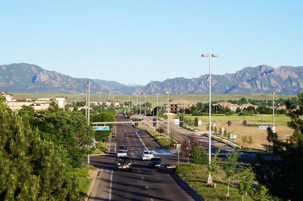 A wide view down a suburban road toward the Flatirons, with cars and traffic lights in the foreground and the jagged rock formations stretching across the horizon under a clear blue sky. Late-day light gives the foothills a soft, warm tone, while trees and new development frame the scene.