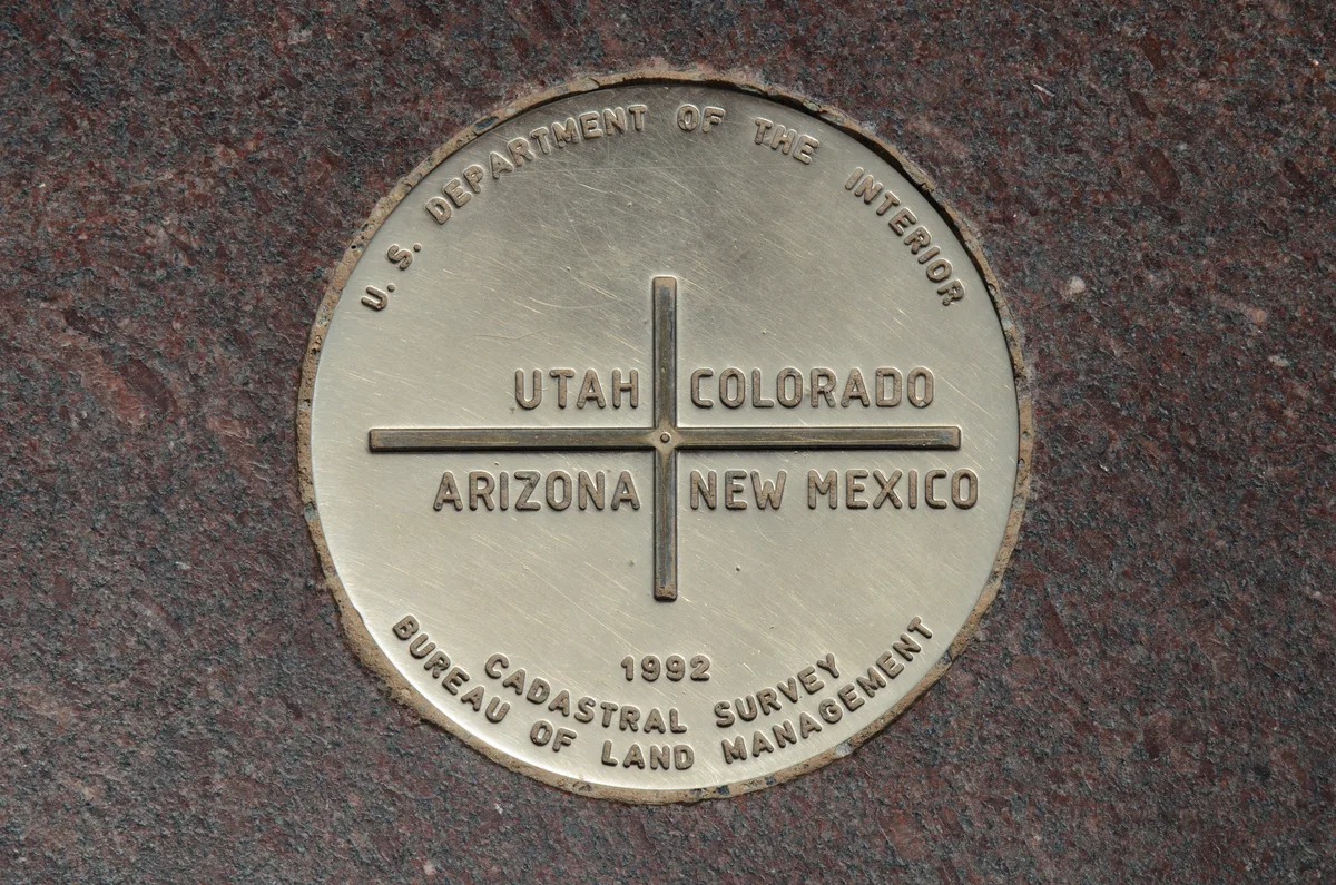 Close-up of the bronze Four Corners marker set in a red granite platform, with a cross dividing the names Utah, Colorado, Arizona, and New Mexico. The warm light brings out the brushed metal texture and the weathered stone around the circular emblem.