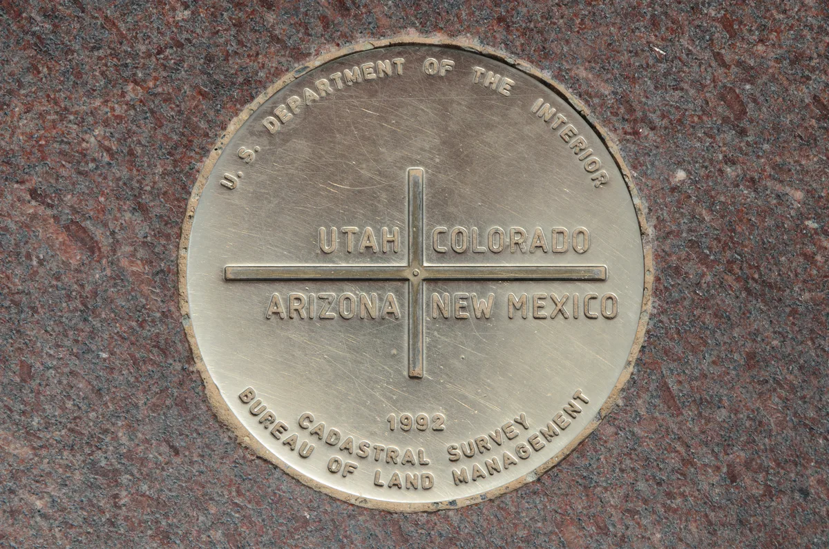 Close-up of the brass Four Corners marker set into red granite, with the names Utah, Colorado, Arizona, and New Mexico divided by a cross at the center. The plaque’s worn metallic surface catches soft light, filling the frame with a tight, symmetrical composition.