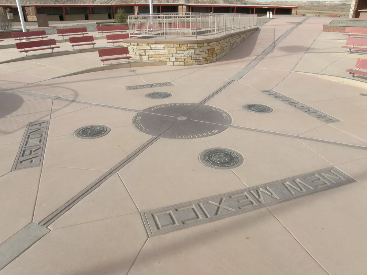 The rebuilt Four Corners marker sits in a sunlit concrete plaza, with bronze state seals and engraved names radiating from the center circle for Colorado, New Mexico, Arizona, and Utah. Red benches and a low stone platform frame the open, high-desert monument area.