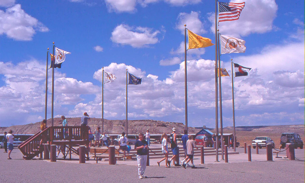 Visitors gather around the Four Corners plaza, where tall flagpoles and state flags rise above the bronze monument area. A bright blue sky with scattered clouds stretches over the open high desert and low mesa horizon.