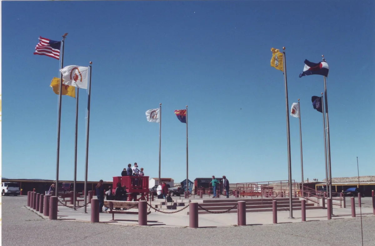 Tall flagpoles ring the Four Corners monument plaza under a clear blue sky, with visitors gathered on the raised platform in the center. The wide concrete and gravel foreground, red railings, and sparse high-desert buildings give the site a stark, open feel.