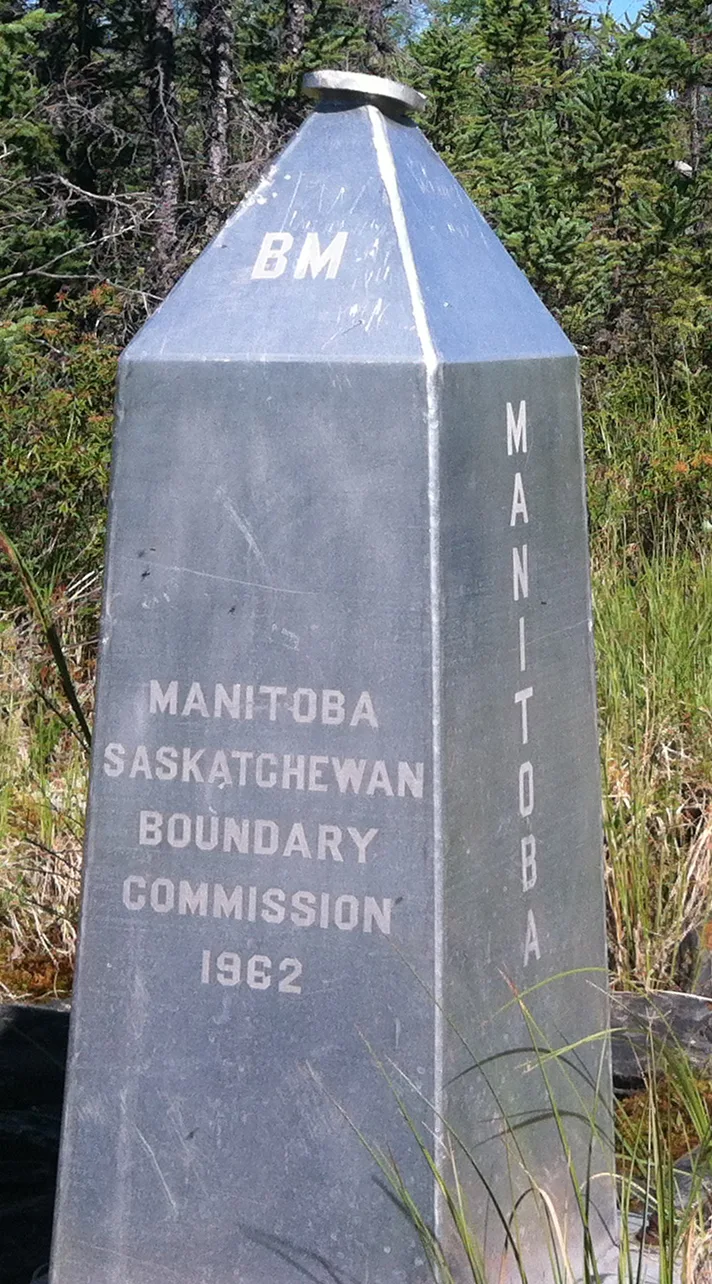A weathered metal boundary marker rises from tall grass, labeled “Manitoba” on one face and “Saskatchewan” on the other, with “Boundary Commission 1962” stamped on the front. Evergreen trees fill the background, giving the monument a remote, forested setting.
