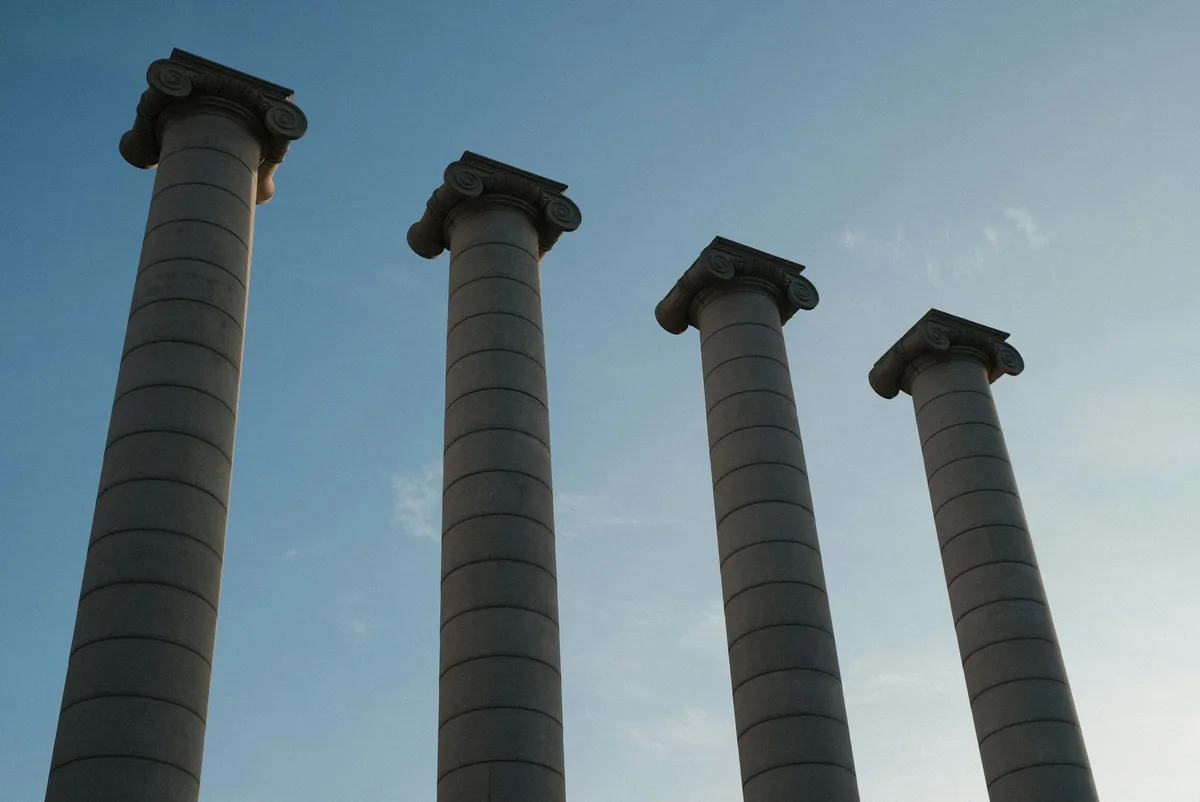 Four tall stone columns with carved capitals rise against a clear blue sky, seen from a low angle that makes them feel monumental. Soft golden light skims the pale columns, emphasizing their rounded shafts and the clean, open composition.