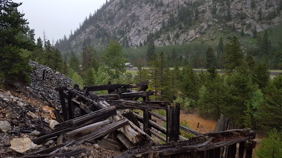 Charred, collapsed mine timbers and a pile of dark rubble sit on a steep, rocky hillside above a road and dense evergreens. A gray mountain face rises behind the scene, partly veiled in mist.
