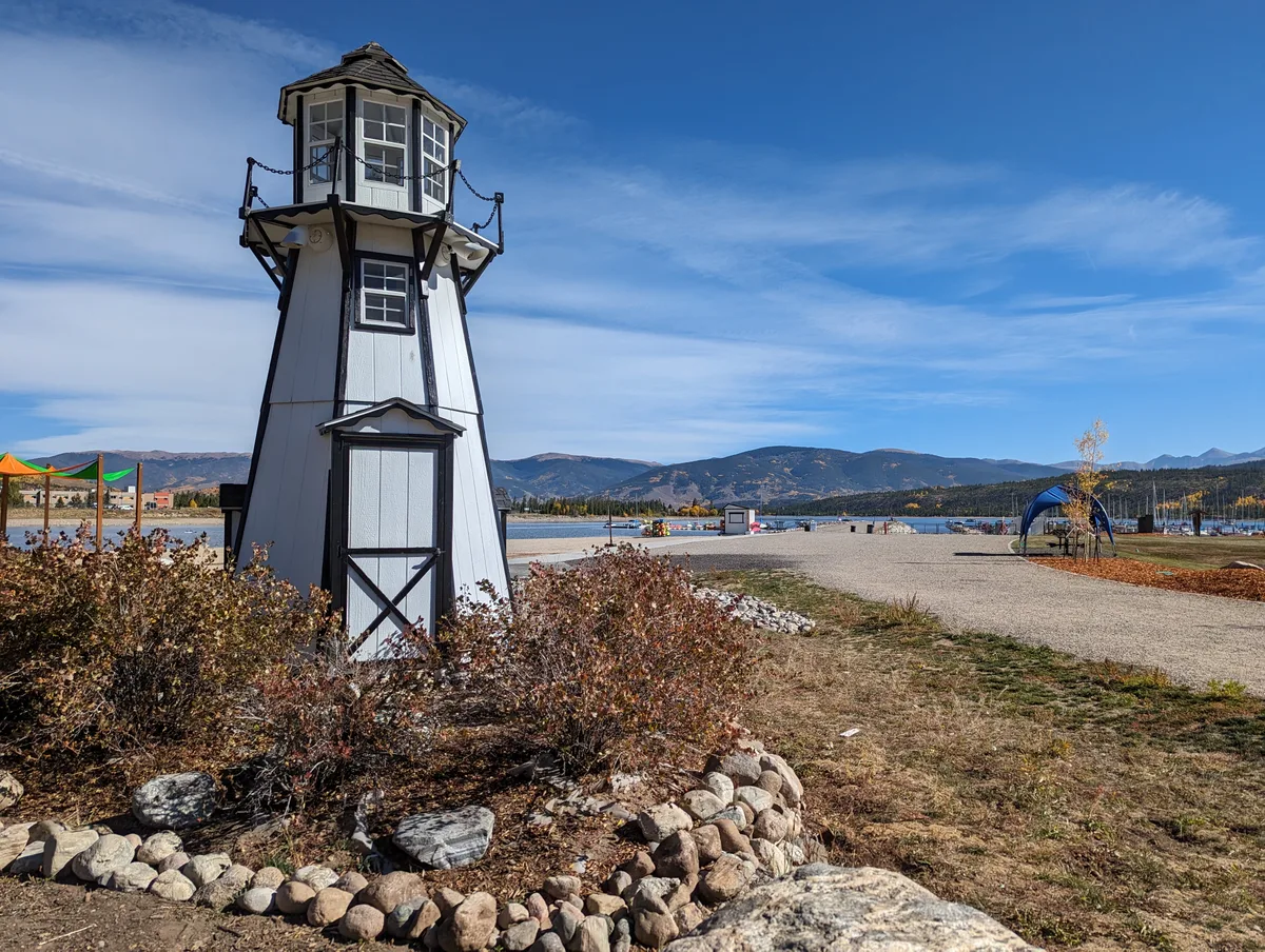 A small white decorative lighthouse stands in the foreground beside a gravel path and low shrubs, with Frisco Bay and the blue-gray Tenmile Range stretching across the background under a clear sky. Scattered marina details, including docked boats and a few colorful shade structures, add interest around the waterfront.