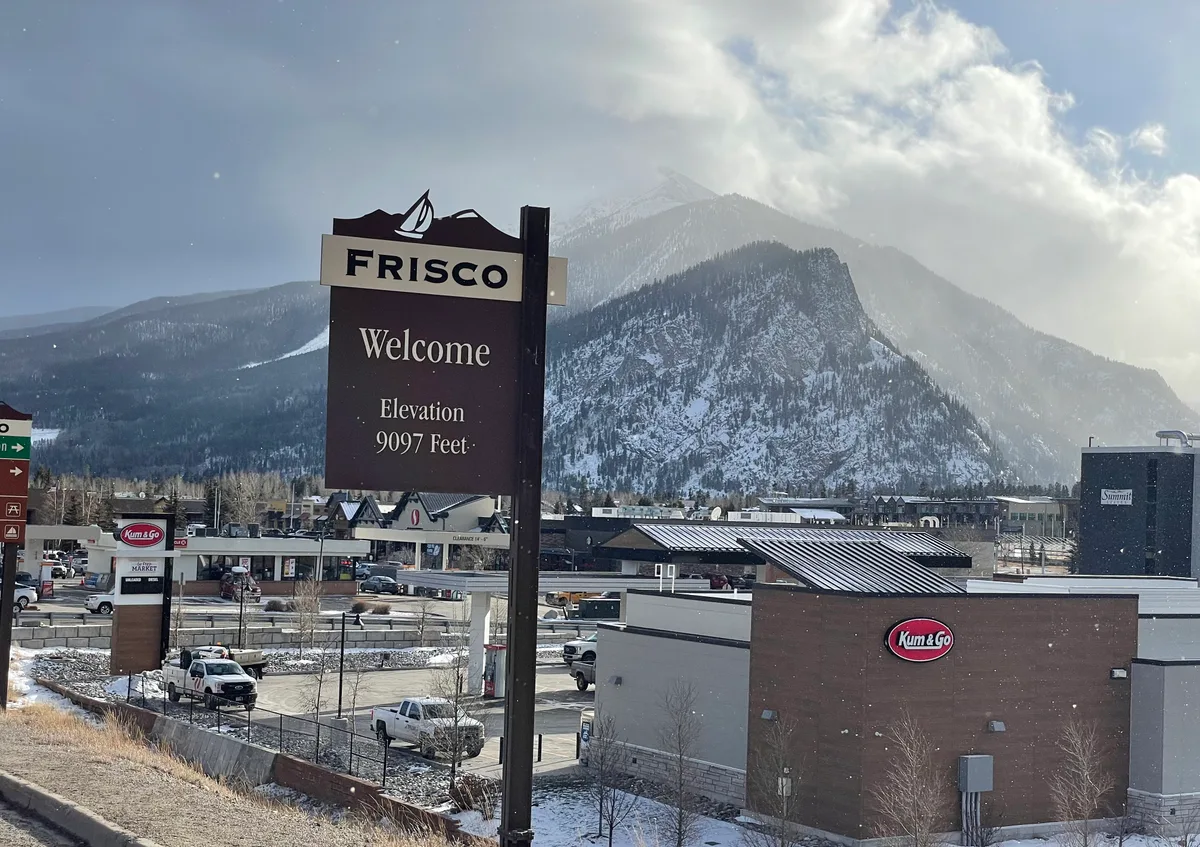 A tall Frisco welcome sign with “Elevation 9097 Feet” stands in the foreground above a snowy town center, with a Kum & Go, parked trucks, and low buildings below. Snow-dusted mountains rise behind the town under bright, hazy winter light and a patchy clouded sky.