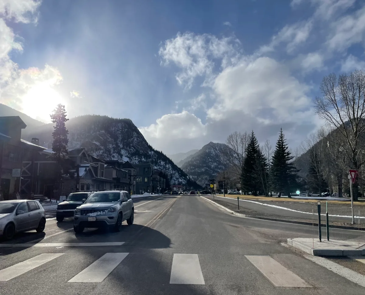 Sunlit mountain town street with a crosswalk in the foreground, parked cars along the left curb, and lodge-style buildings lining the road. Snow-dusted peaks rise in the distance under a bright blue sky with scattered clouds and a low sun glowing at the left edge.