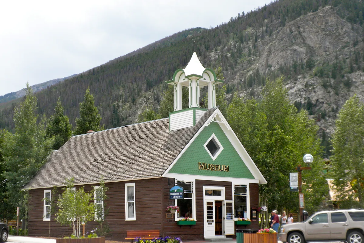 A small historic museum with a steep shingled roof and white bell tower sits at the base of a rocky, pine-covered mountain, framed by summer trees. The green gable front and flower boxes add color, while parked vehicles and a few people line the quiet street in front.