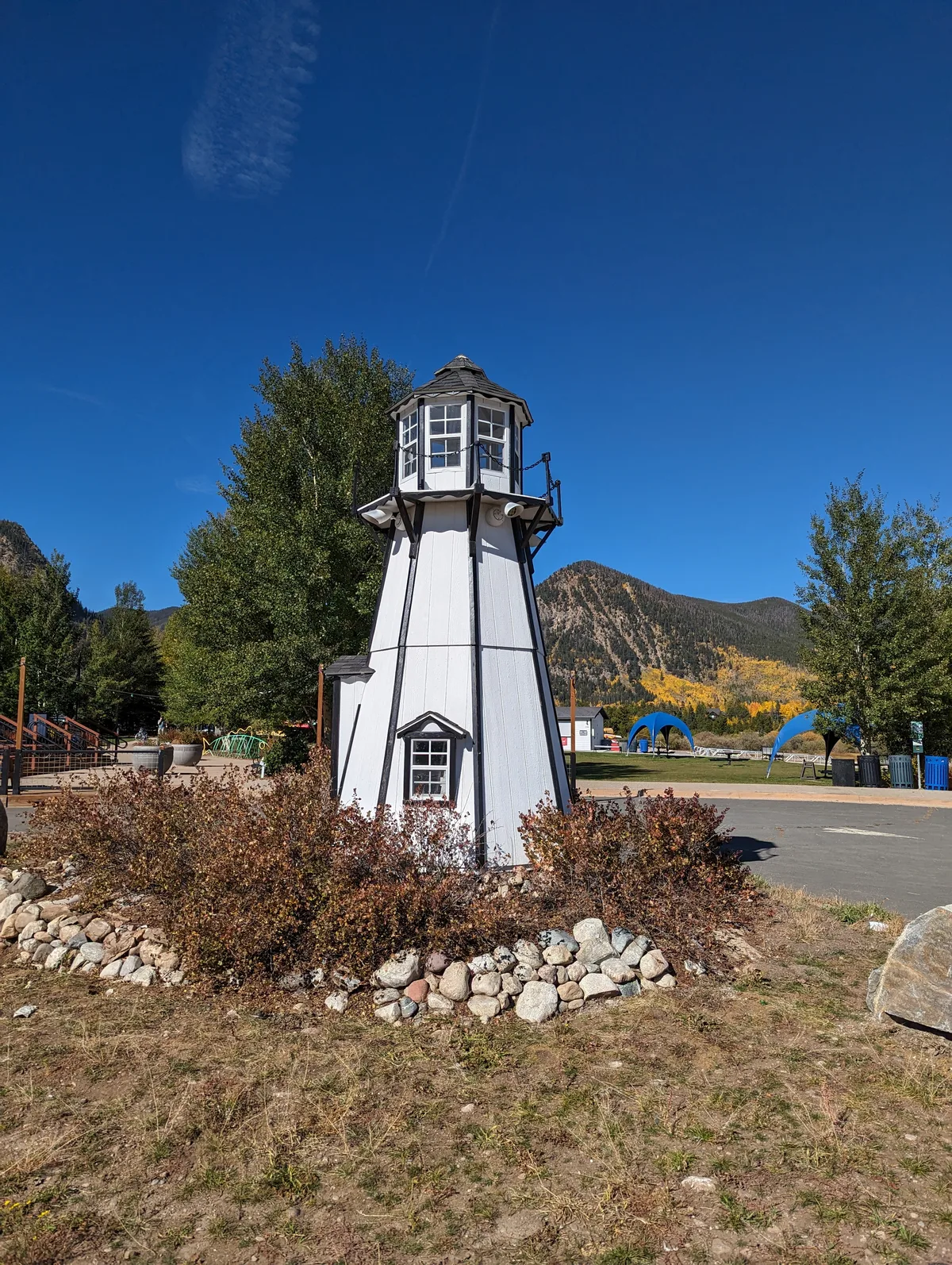 A small white lighthouse-style structure stands in the marina, ringed by rocks and autumn shrubs, with bright blue sky overhead. Behind it, the Tenmile Range rises above trees dotted with yellow fall color and a few blue shade canopies on the waterfront lawn.