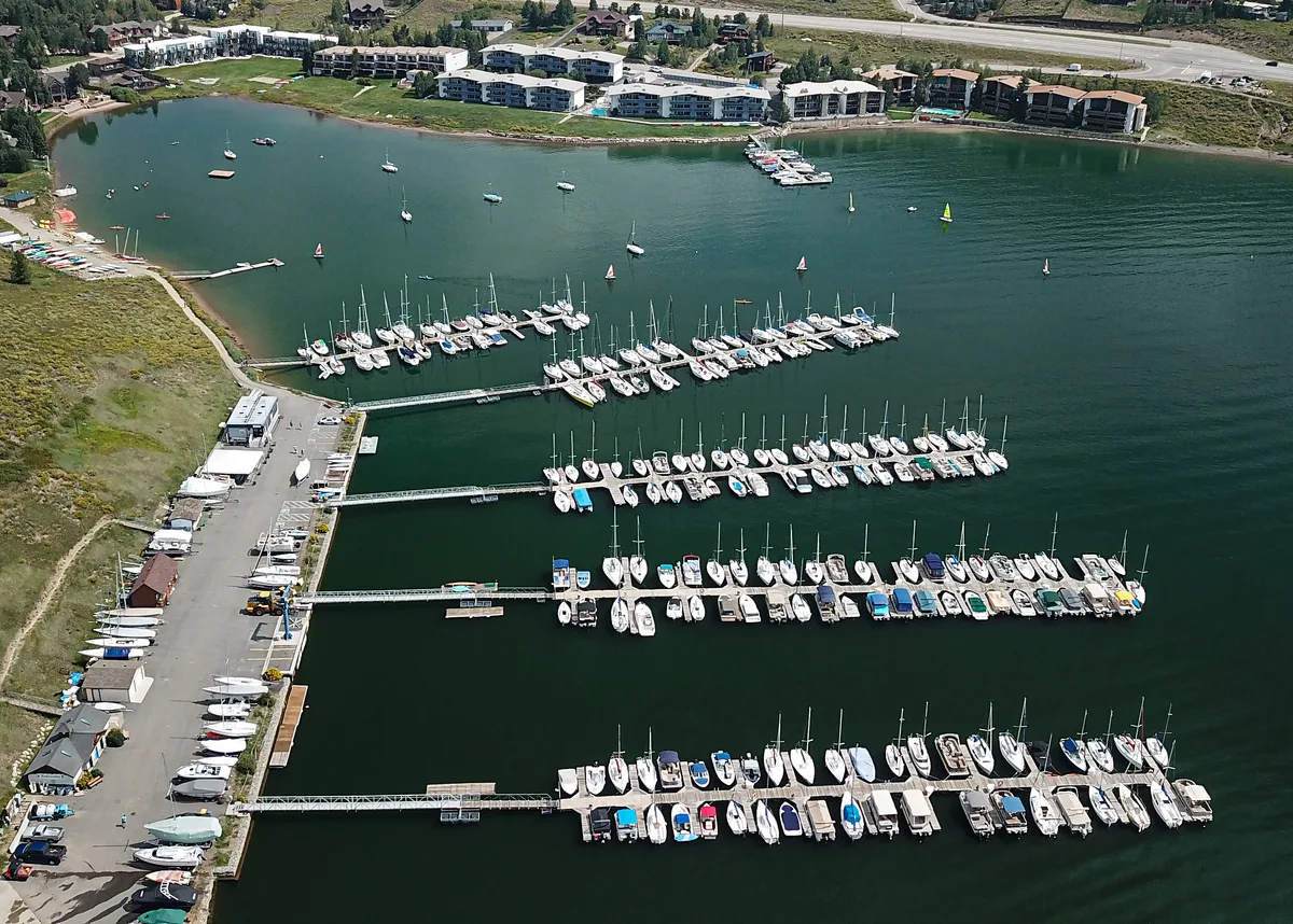 High-angle view of Frisco Bay Marina with several long docks packed with white sailboats and a few small colored boats scattered across the dark green reservoir. The shoreline parking area, boatyard, and low lakeside buildings line the left edge, with calm water and soft golden-hour light giving the scene a quiet, orderly feel.