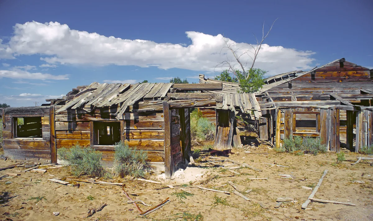 Weathered wooden ruins of a long-abandoned building sit in a sandy, scrubby landscape, with broken roof beams, empty window frames, and scattered debris in the foreground. Bright sunlight and a deep blue sky with a broad white cloud add sharp contrast to the crumbling structure.