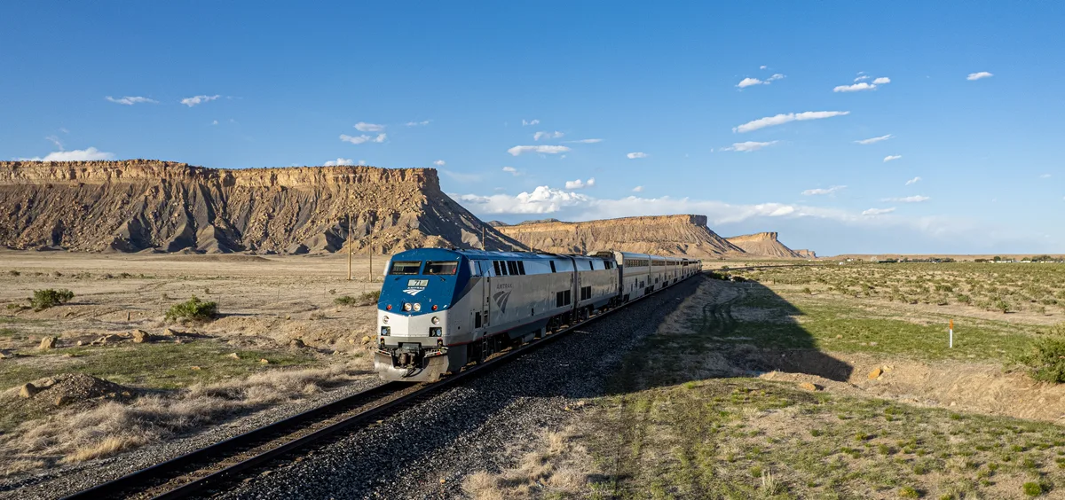 A silver California Zephyr train curves through the high desert foreground, heading toward the layered Book Cliffs under a bright blue sky. Warm golden light rakes across the cliff faces, while long shadows stretch over the sparse grassland and rail bed.