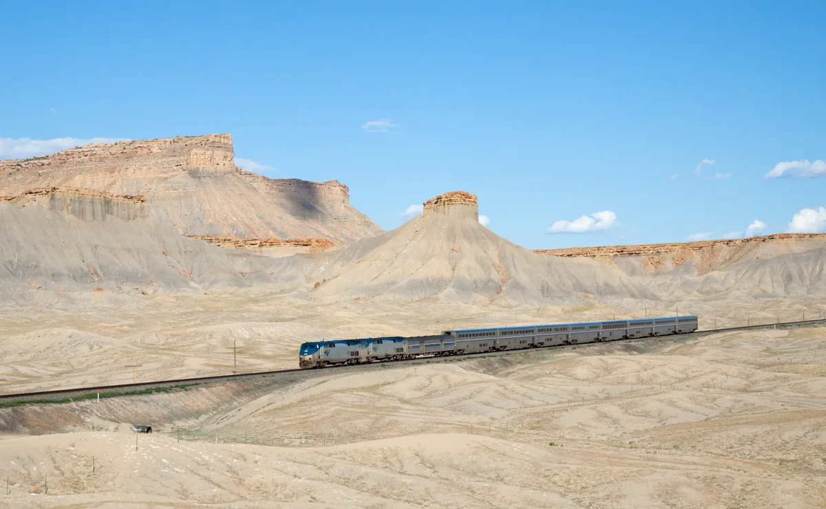 A long passenger train winds across a pale desert plain below towering, layered Book Cliffs and a rounded butte, all under a clear blue sky. Soft golden light highlights the tan and gray rock faces while the sparse foreground shows dry, striated badlands.