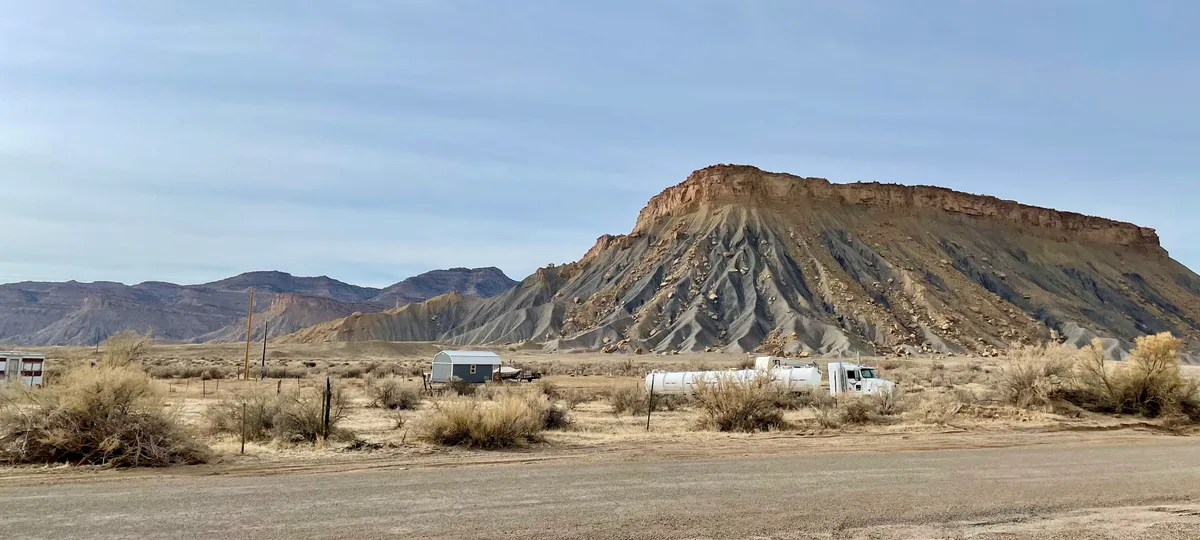 A wide desert view shows the layered Book Cliffs rising steeply behind a flat, scrubby plain, with dark gray gullies and a rust-colored caprock glowing in soft golden light. A few small buildings, a parked truck, and a road in the foreground emphasize the scale of the escarpment.