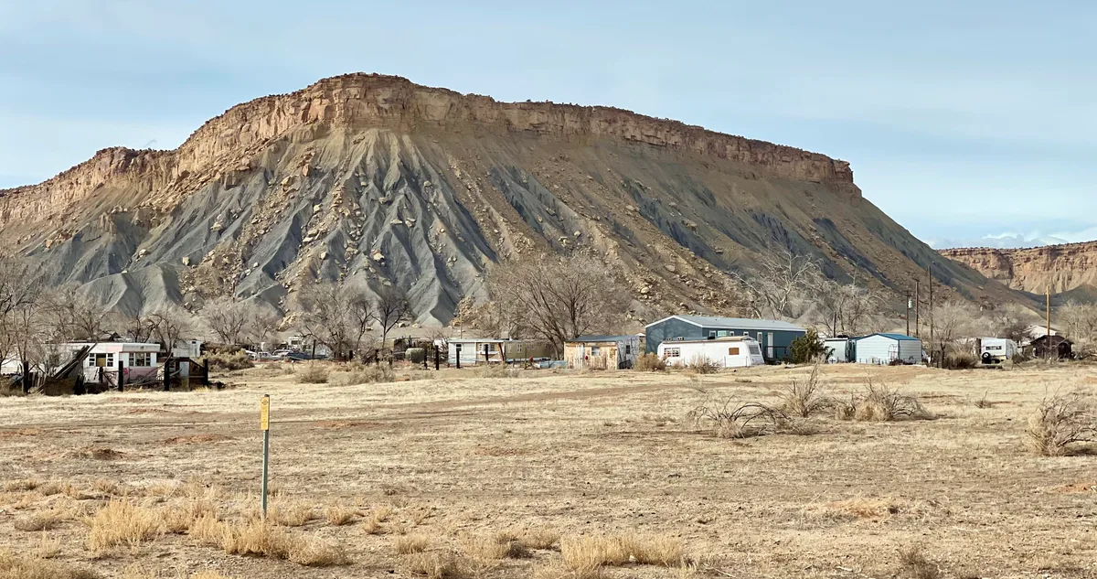 A broad desert valley stretches toward the towering Book Cliffs, with tan and gray layered rock rising above a few small homes and leafless trees. Soft golden light warms the cliff top while the lower slopes sit in cool shadow under a pale blue sky.