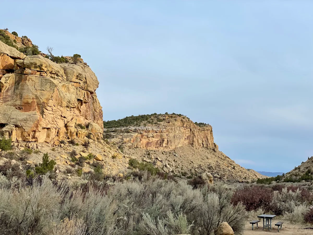 Golden light warms layered sandstone cliffs rising above a scrubby desert valley, with a pale blue sky overhead. Low shrubs and a small picnic table in the foreground add scale to the wide, rugged landscape.