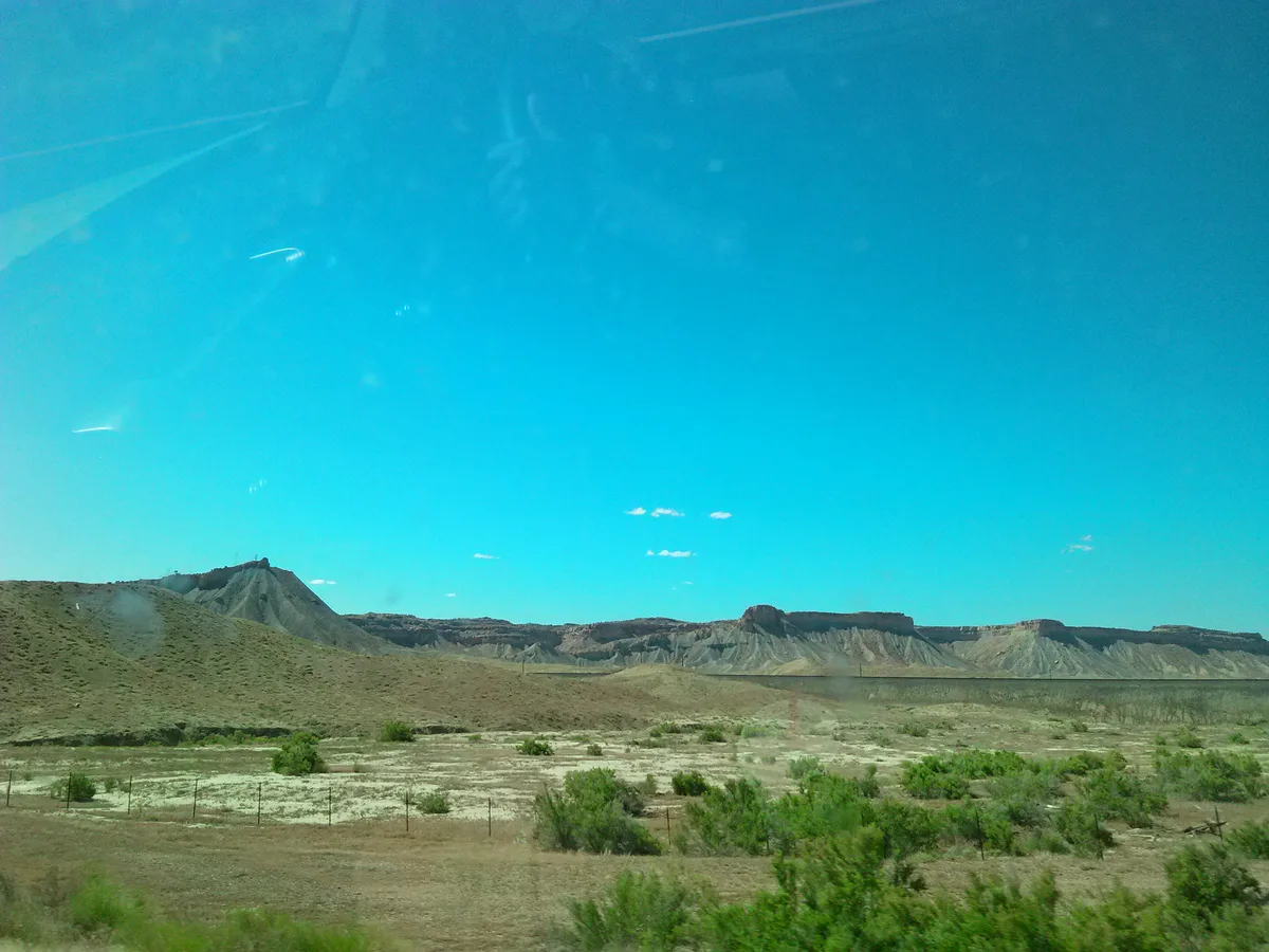 Wide view across a dry high-desert plain with scattered green shrubs, leading to the long, layered Book Cliffs stretching across the horizon. The cliffs sit in hazy afternoon light beneath a vivid blue sky, with pale tan slopes and darker mesas marking the escarpment.