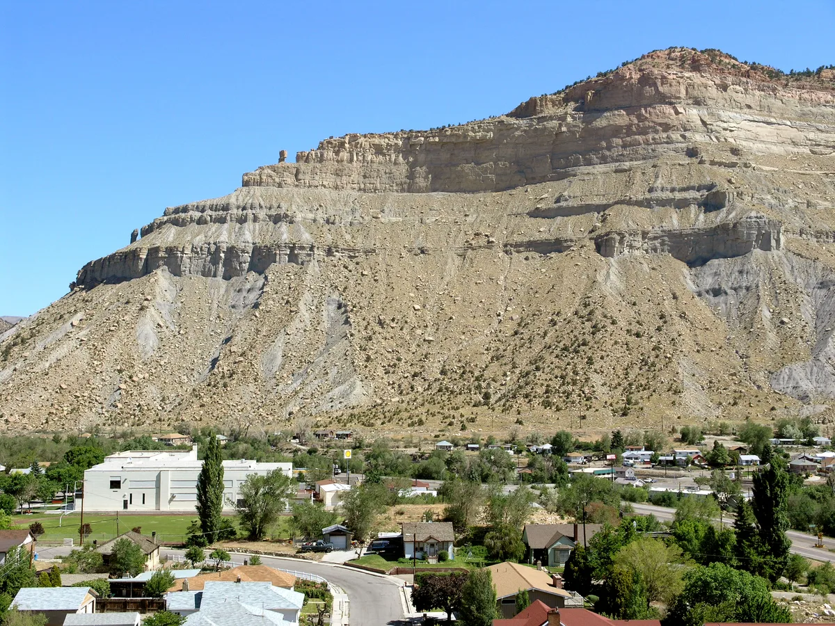 A massive layered sandstone cliff rises above a small desert town, with pale tan and gray rock bands stepping up toward a flat ridge under a clear blue sky. Houses, trees, and a curving road fill the foreground, emphasizing the scale of the Book Cliffs backdrop.
