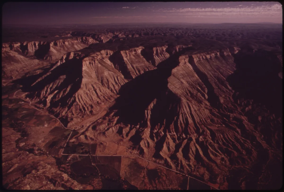 Wide view of the Book Cliffs rising in rugged, layered ridges above the Grand Valley, with long purple shadows cutting across the desert slopes. Golden light warms the cliff faces under a hazy sky, emphasizing the steep escarpment and dry high-desert terrain.