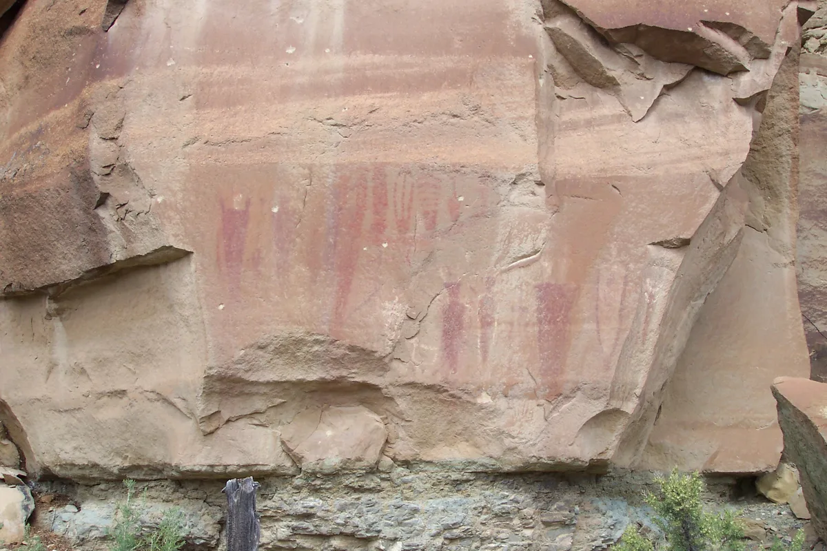 A close view of a sandstone cliff face shows faded red pictographs painted on a pale, weathered rock wall with broken, angular layers. Sparse desert vegetation grows at the base beneath the overhanging cliff.