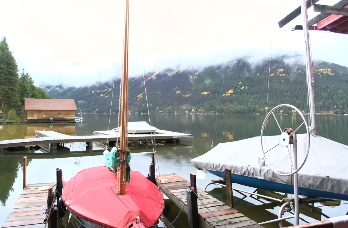 Boats covered with red and white tarps sit in a wooden marina on still Grand Lake, with docks leading into the glassy water. Low fog hangs over the dark forested mountains across the lake, and patches of golden sunlight catch the slope above the shoreline.