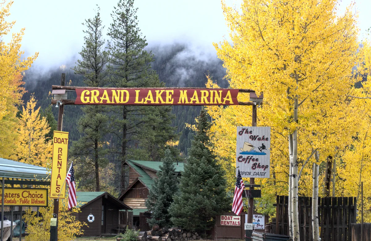 The entrance to Grand Lake Marina is framed by bright golden autumn aspens and dark evergreen pines, with misty mountain slopes fading in the background. A red-and-gold marina sign, boat rentals, and small rustic buildings with American flags give the scene a cozy western-lake feel.