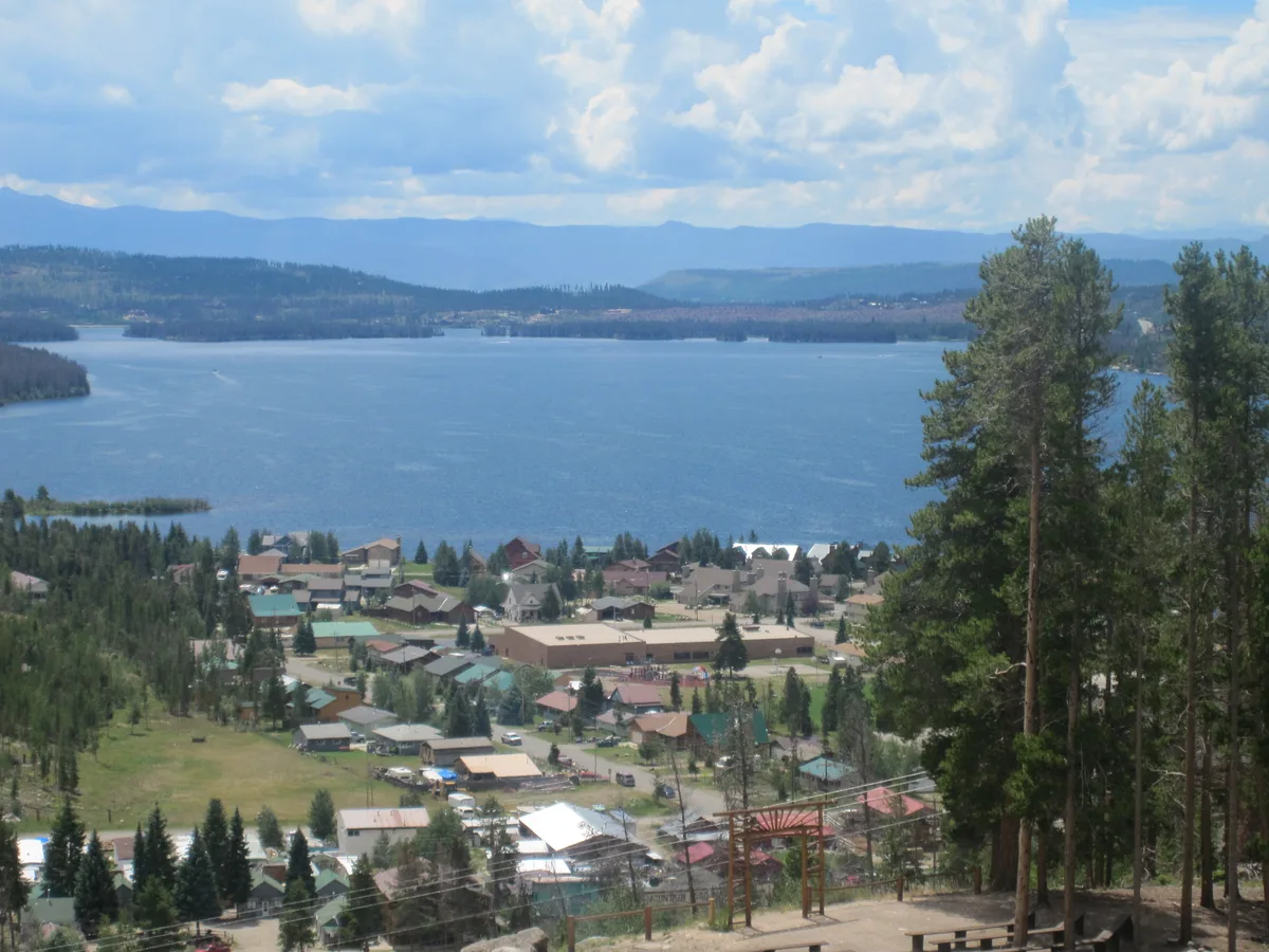 A wide view looks down over Grand Lake and the town below, with blue water filling the middle of the frame and low mountain ridges fading into the distance under a partly cloudy sky. Tall pines in the foreground frame the scene, adding depth to the lakeside landscape.