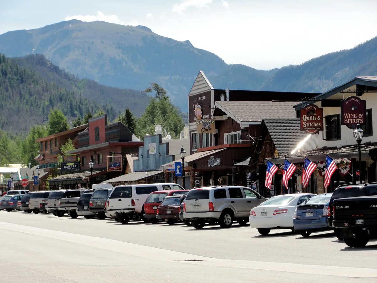 Downtown Grand Lake’s main street is lined with parked cars, storefronts, and American flags in bright sunlight, with steep forested mountains rising behind the town. A hazy blue ridgeline and high-altitude sky frame the western slope setting.