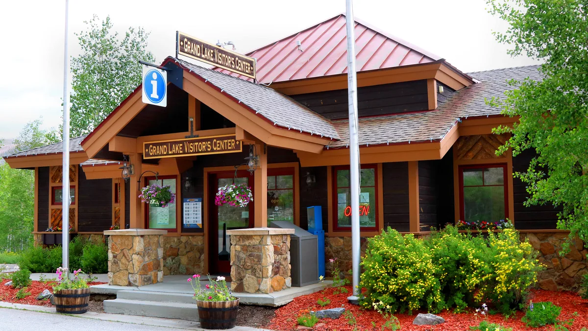 The Grand Lake Visitor’s Center sits in a rustic lodge-style building with a red metal roof, stone porch columns, and dark wood siding, framed by leafy trees. Bright flower baskets, red mulch, and an illuminated “OPEN” sign add color to the front entrance.