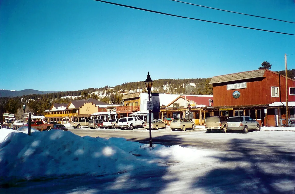 Snow-covered Main Street in Grand Lake with parked cars, rustic storefronts, and the “Boot Hill” building in warm golden light. A clear blue sky hangs over the small mountain town, with forested hills and patches of snow on the slopes in the background.