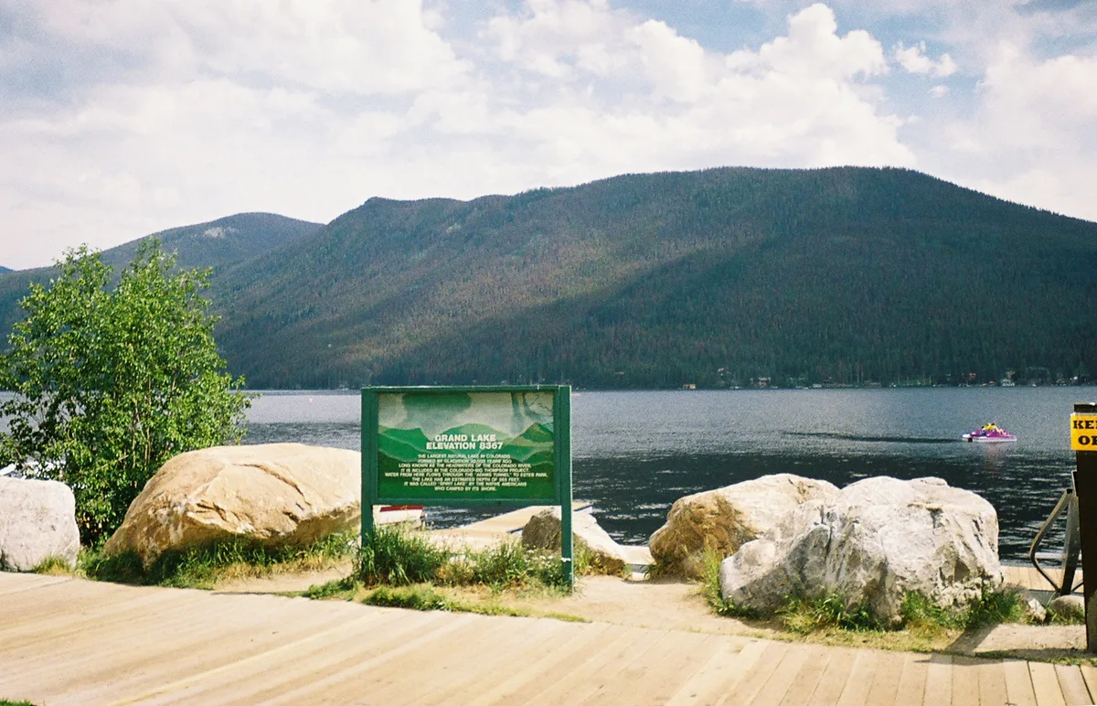 Grand Lake stretches across the frame beneath dark, forested mountains, with soft clouds overhead and a calm, reflective surface. In the foreground, a green Grand Lake sign sits between large boulders on a wooden overlook, with a small boat visible near the far shore.